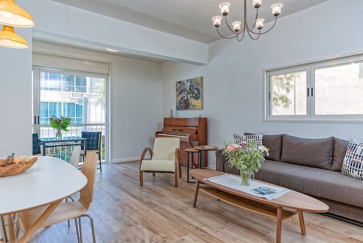 Bright living room in a pilot holiday apartment, with a sofa, flowers and a piano beside the balcony.